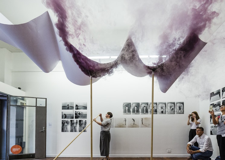 A femme-presenting figure in a striped shirt and khaki green pants stands under a banner structure from which purple coloured smoke emanates