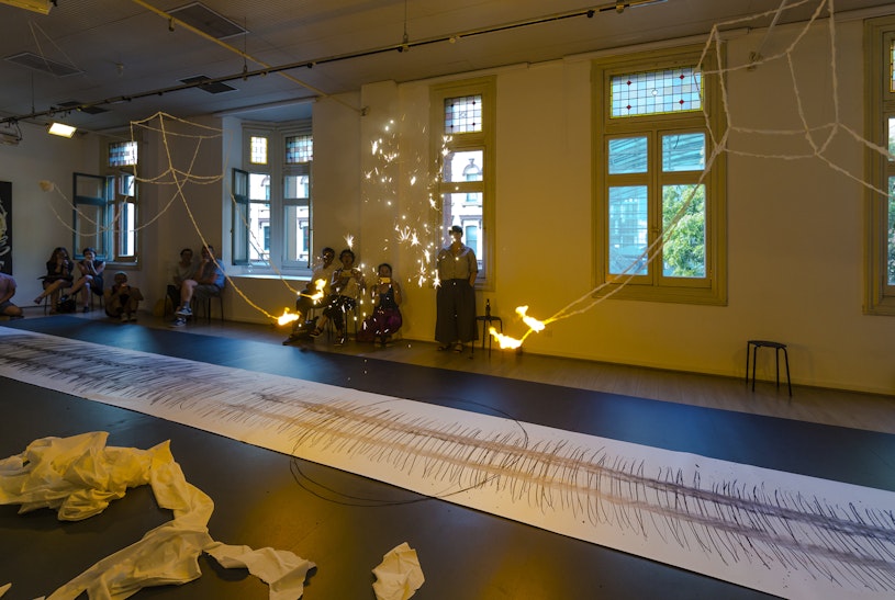 Bright sparks explode in a dim room between four lines of burning rope, over a long scroll of white paper. Seated visitors look on from each side of the scroll