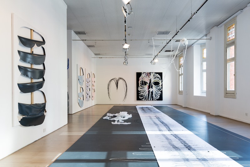 A white gallery space with a long strip of white paper covered in charcoal marks, large canvases that have been cut or scribbled over, and curved black lines on the wall