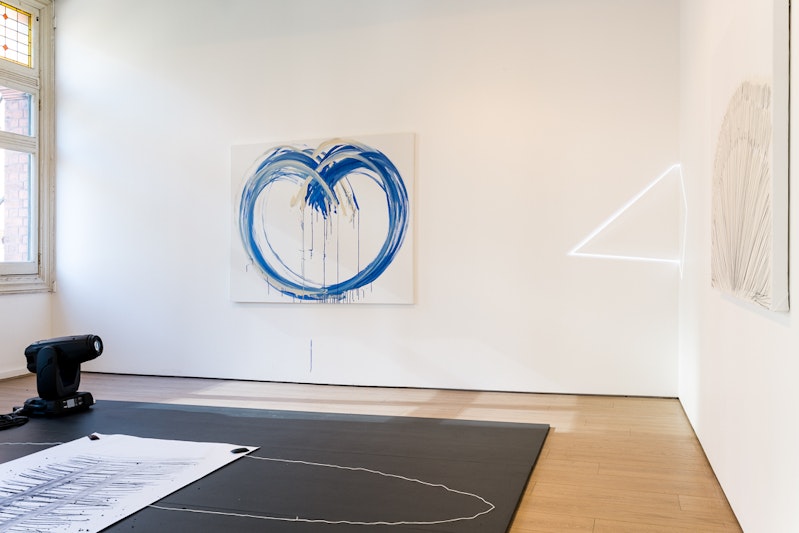 Blue and cream-coloured dripping lines painted on a white canvas, with light beams curving around one corner of a white gallery space. On the floor is a black landing with a strip of white paper covered in charcoal lines