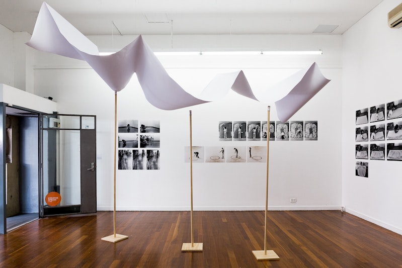 A white gallery space with a banner structure folded over three wooden poles. On the walls are black and white prints of an artist drawing on the ground with charcoal and scribbling on a wall in white