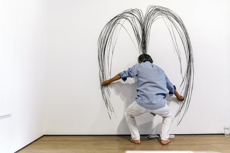 A silver-haired figure in a blue striped shirt and white paints stands with bent knees in front of a white wall while scribbling curved lines with a stick of charcoal in each hand