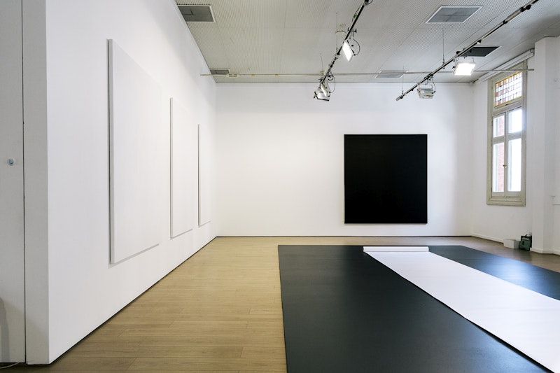 A white gallery space, with three white canvases hanging on the left wall, a black canvas hanging on the back wall, and a black landing on the floor with a long strip of white paper rolled out on top