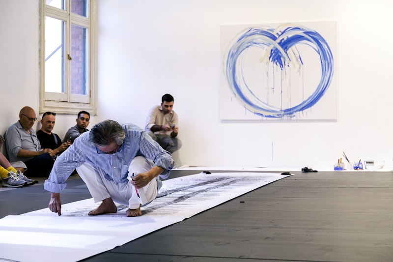 A male-presenting figure with silver hair, a blue striped shirt and white paints squats barefoot on a long scroll of white paper, drawing with a stick of charcoal