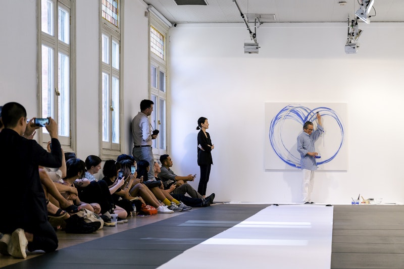 An East Asian male-presenting figure in glasses and a blue shirt paints blue circles on a white canvas. A crowd sitting and standing against a white wall look at him and take photos