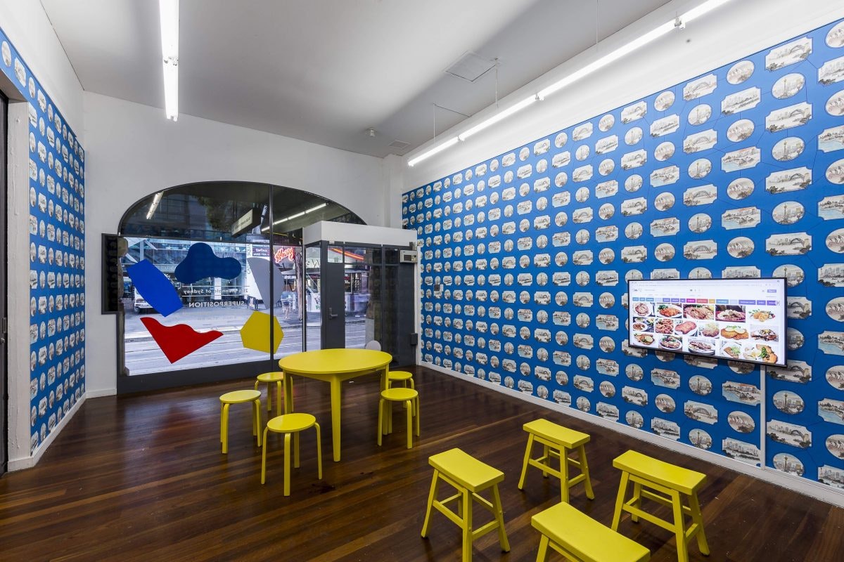 A gallery space with blue wallpaper printed with vignettes of the Sydney city skyline, and a television screen mounted on the wall. The glass window is has a print of colourful irregular decal shapes stuck at the front. There is a round yellow table surrounded by five round yellow stools, and four rectangular stools arranged in front of the television screen.