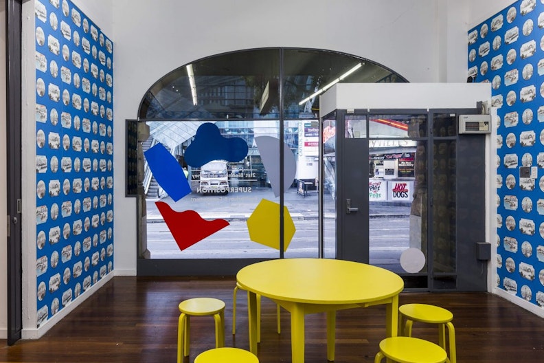 A round yellow dining table surrounded by round yellow stools in a gallery space decorated with blue wallpaper.