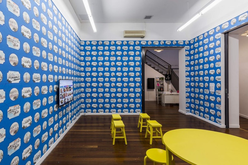 Four yellow rectangular stools arranged in a gallery space with blue wallpapered walls.
