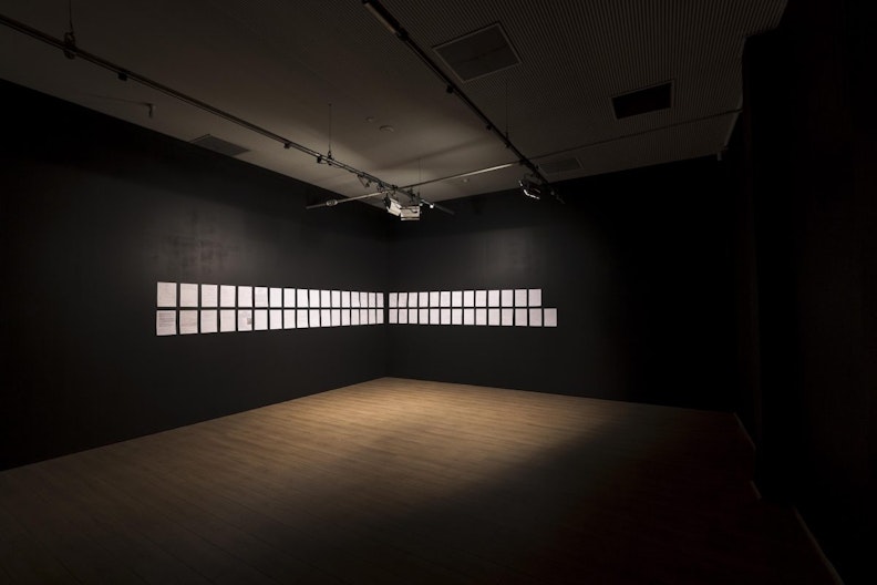 A gallery space with two rows of white A4 paper fixed on two black gallery walls. They are spotlit by some gallery lights.