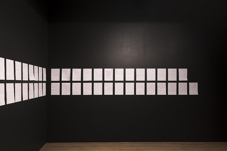Two rows of white sheets of A4 paper printed with text, mounted on a black gallery wall.