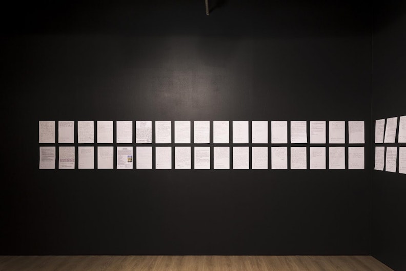 Two rows of white sheets of A4 paper printed with text, mounted on a black gallery wall.