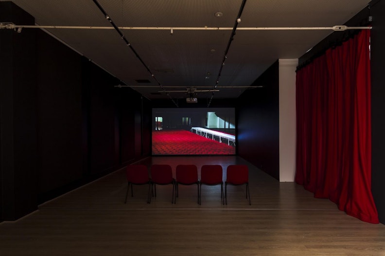 A row of five red fabric chairs arranged in a dark room, in front of a screen with a video projection. On the left is a black gallery wall, on the right is a long red velvety curtain.