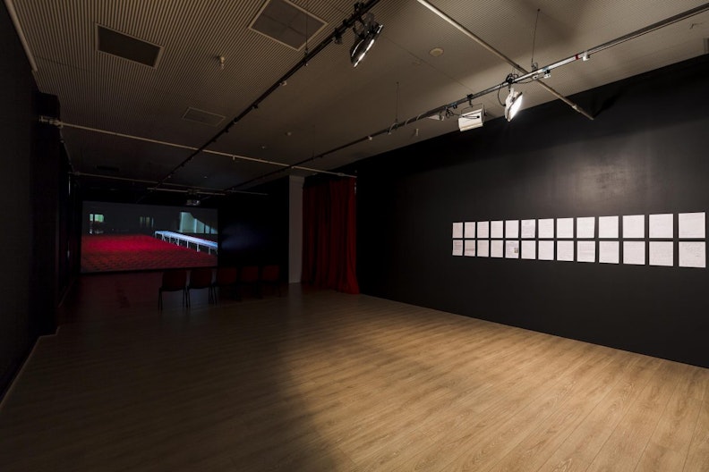 A gallery space with black walls and a red velvety curtain. On furthermost wall is a video projection of a red floor. On the right of the gallery space is a series of white A4 sheets mounted on the wall under a gallery spotlight.