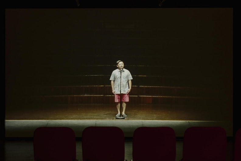 A video still of a male-presenting figure of East Asian appearance standing at a stand-up microphone on a dark, empty stage. He wears glasses, a button up short-sleeved shirt and salmon pink shorts, with slipper-like shoes. His arms are resting by his sides.