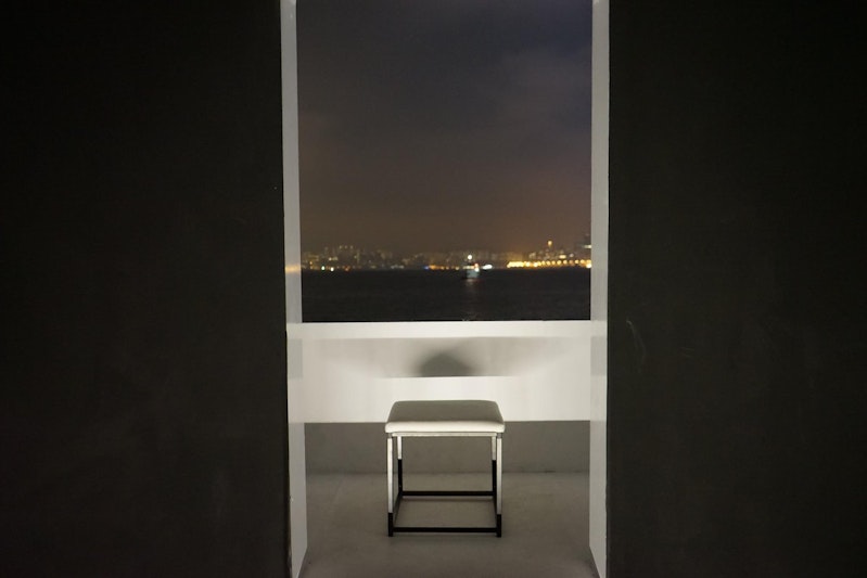 A white rectangular stool on a white balcony that looks across a harbour at night with buildings and city lights on the other side.