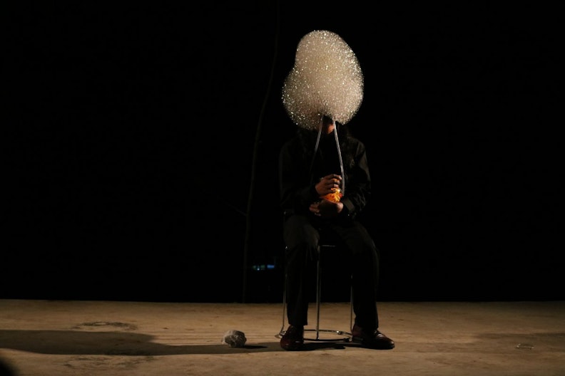 A figure in black sitting on a stool against a black background, with a cloud of bubbles obscuring their face.