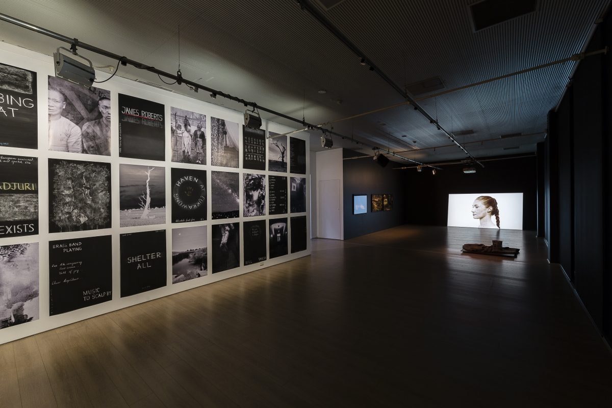 A dim-lit gallery space with a series of black and white posters on the left wall and a video of a female face with red hair braided back projected onto the back wall.