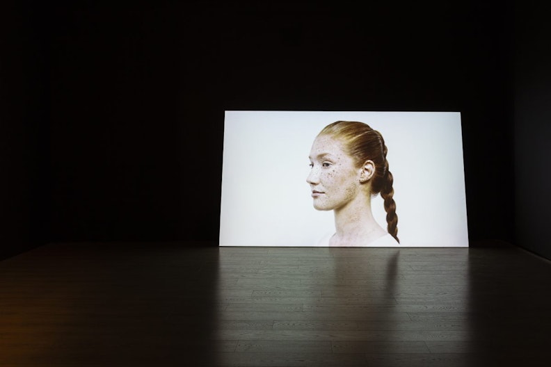 A dark gallery space with a projection of a Caucasian woman's face. She has red hair braided back, freckles along her checkones and along her jawline and neck.