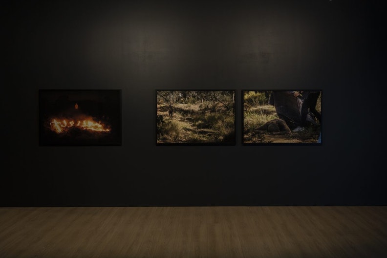 Three framed photographic prints on a grey gallery wall. The left shows a figure watching a series of the fires in the night, the middle print is of some grassy riverbeds in the Australian outback and the right shows a figure lying facedown in the dirt by the river, with a second figure shown legs-down pulling a covering over the lying figure.