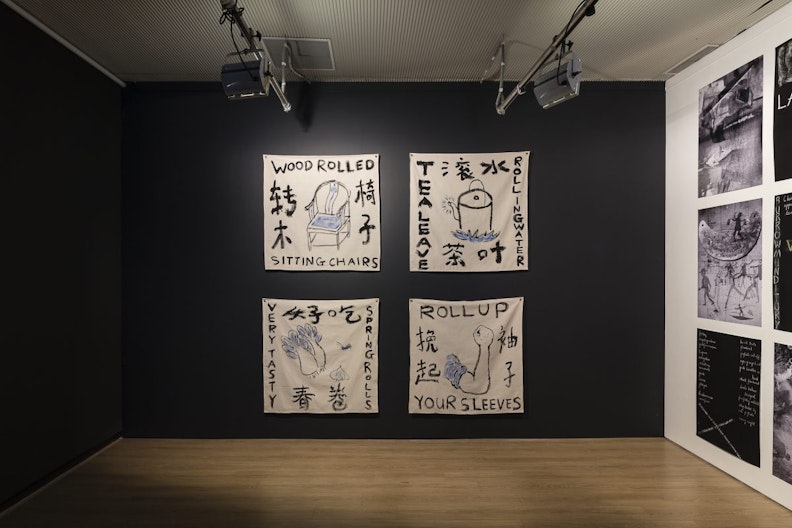 Four white cotton sheets painted with symbols, English words and Chinese characters. The upper left sheet shows a chair painted in black and blue ink, surrounded by Chinese characters and the words 'WOOD ROLLED SITTING CHAIRS'. The upper right sheet shows a painted teapot boiling on a blue flame, surrounded by Chinese characters and the words 'TEA LEAVE ROLLING WATER'. The lower left sheet shows two bok choy, a garlic head and an insect surrounded by Chinese characters and the words, 'VERY TASTY SPRING ROLLS'. The lower right sheet shows a curled bicep and fist with a blue rolled up sleeve, surrounded by Chinese characters and the words, 'ROLL UP YOUR SLEEVES'.