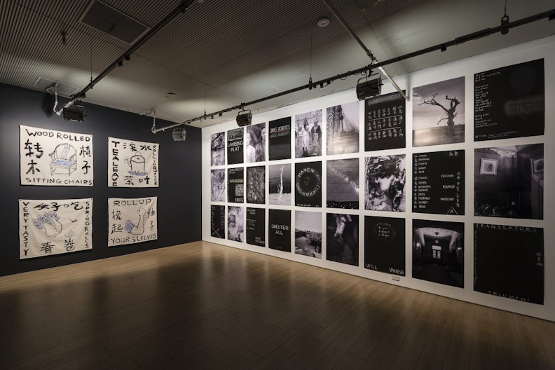 A dim-lit gallery space with four white cotton sheets painted with symbols, Chinese characters and English words in black and blue ink on the left wall. On the right wall is a series of black and white posters, showing English and Chinese names, barren trees in the Australian outback and figures of East Asian appearance.