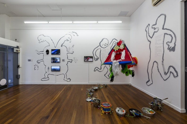 A white gallery space with three dancing characters painted on the white walls. One of the figures has two heads situated on two long necks, four arms and two feet, with three LED screens lined straight down the body. A ring of stainless steel pots, kitchen utensils and toy drums is arranged on the hardwood floor.