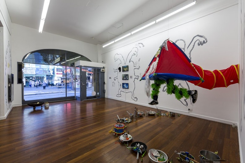 A red kid's play tent is suspended from a white gallery ceiling and attached to the wall with a matching play tunnel. Two mannequin legs wrapped in two feathery green boas hang from the blue floor of the tent. Black sneakers are attached to the ends of these legs. The tent is suspended over a ring of stainless steel pots, cooking utensils and a toy drum.