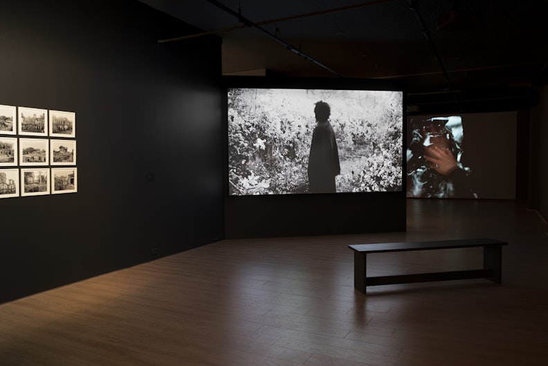 A dark room with black walls with a set of black and white photographic prints framed in white on the left, and video projections on two screens set up in front of a black bench.