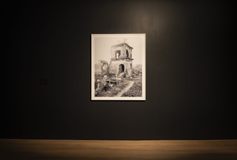 A black gallery wall with a spotlight photo of a grassy overgrown path leading up to a ruined building.