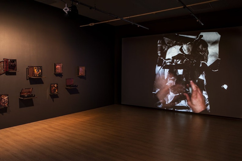 A video projection showing a pair of hands crumpling and tearing up a printed photograph. On the left is a series of wooden boxes fixed to a black gallery wall.