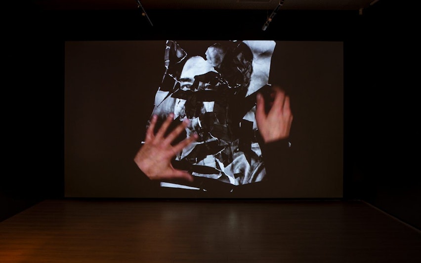 A video projection showing a pair of hands in motion, crumpling and tearing up a large black and white photograph.