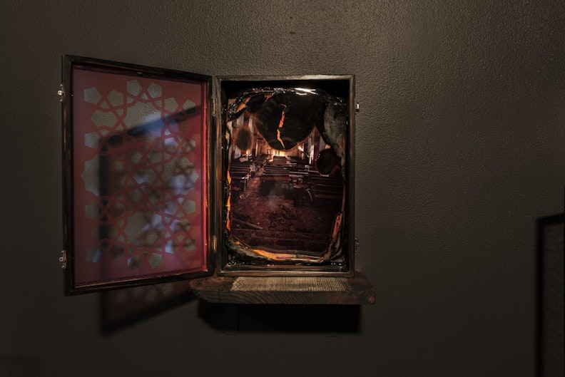 A photograph with burnt edges set in an open wooden box hung on a black gallery wall. The photograph shows rows of pews.