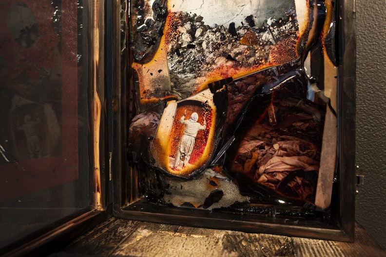 Close-up of a photograph set in an open wooden box. The photograph has burnt edges and appears to be a collage of images, showing rubble and a hooded figure throwing their hands up in the air.