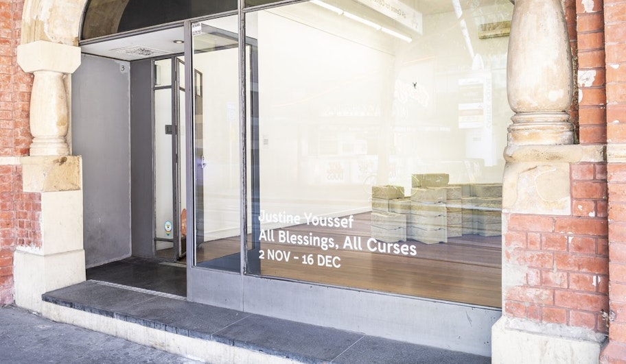 A glass facade in a red brick building, with the decal words 'Justine Youssef, All Blessings, All Curses, 2 Nov - 16 Dec' stuck on the glass.