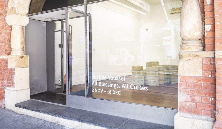 A glass facade in a red brick building, with the decal words 'Justine Youssef, All Blessings, All Curses, 2 Nov - 16 Dec' stuck on the glass.