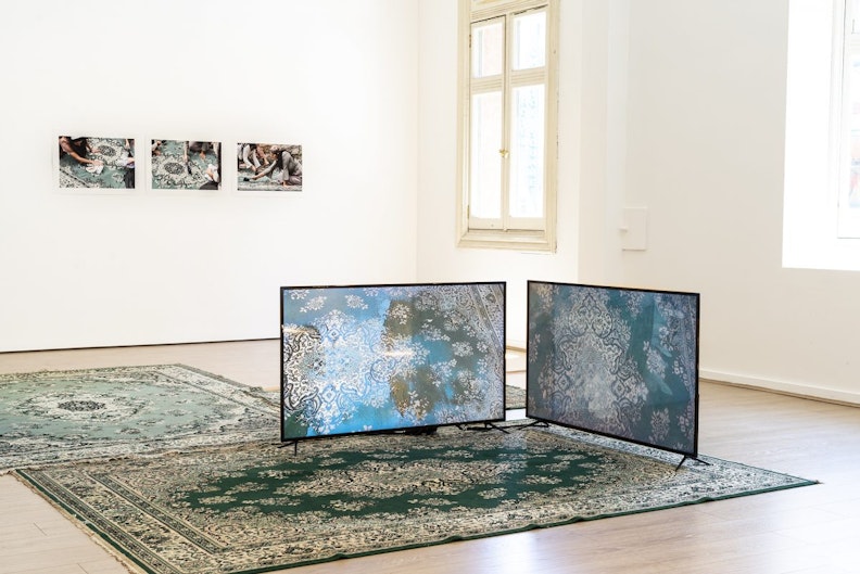Two teal green Persian rugs laid on a hardwood floor with two LED video screens stood on top, showing the same displayed rugs in other settings.
