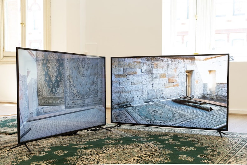 Two LED video screens on a teal green Persian rug. The screens show a series of Persian rugs laid out in other concreted spaces.