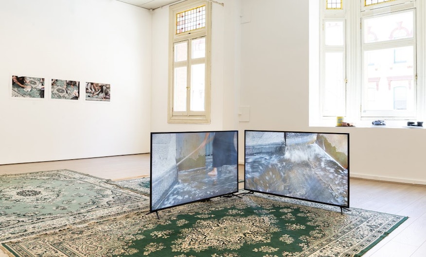 Two green-toned Persian rugs in a light-flooded gallery space, with two LED screens stood on top showing a woman legs-down cleaning a floor.