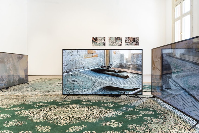 A teal green Persian rug with an LED screen on top showing a series of folded Persian rugs in a tiled floor in a concrete building.