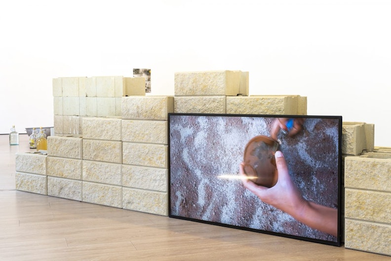 A wall constructed from sandstone brick with a LED video screen showing two tanned hands tipping a clay bowl over onto the ground.
