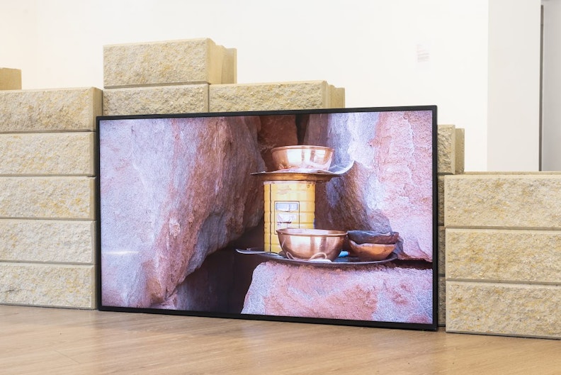 An LED video screen showing three brass bowls and a yellow tin can on a natural red rock shelf.