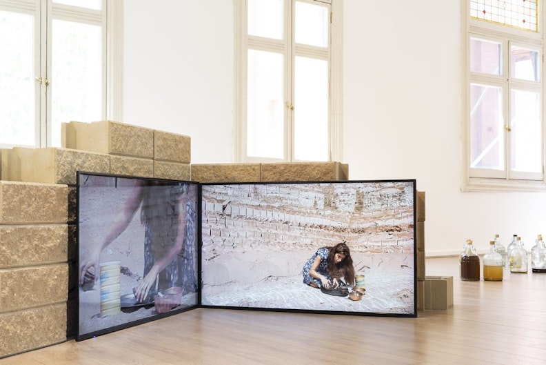 Two LED video screens showing a femme-presenting woman with long brown hair in a desert landscape leaning over a set of brass bowls and a yellow tin can.