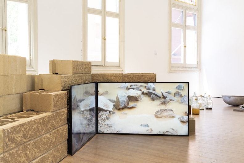 Two LED video screens against a small sandstone brick wall, showing a video of granite rocks in brown water.