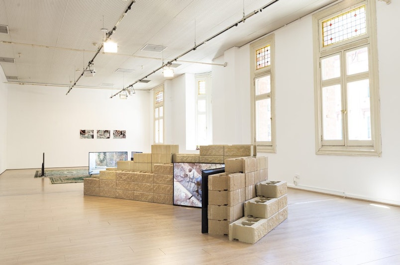 A small brick sandstone wall, several LED TV screens and two teal green Persian rugs arranged in a white gallery space.