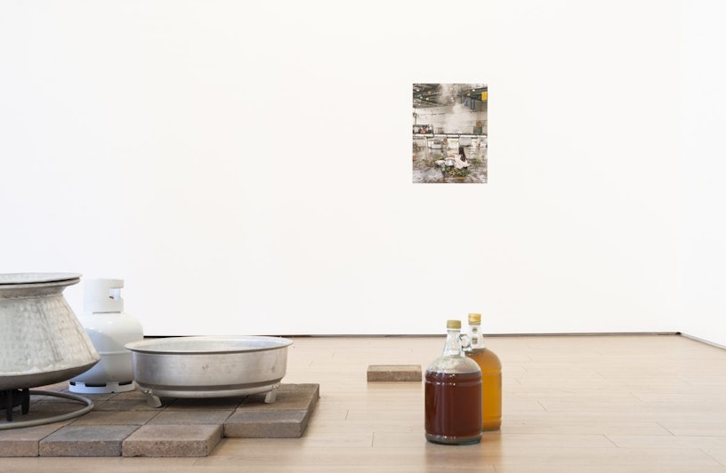 A two ring gas burner, gas cylinder, aluminium pot on a pavers floor, next to two glass bottles filled with brownish liquids. On the white wall behind are two photographs.