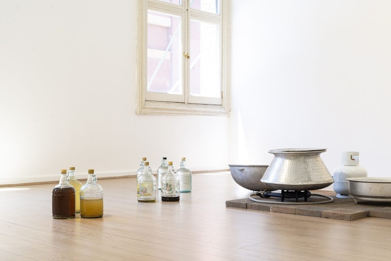 Light coming through a gallery window onto eight glass bottles filled with different liquids, arranged next to an aluminium pot, a two ring gas burner and a gas cylinder on a pavers floor.