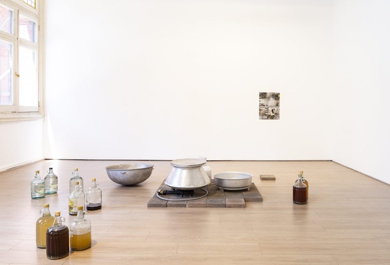 Light coming through a gallery window onto eight glass bottles filled with different liquids, arranged next to an aluminium pot, a two ring gas burner and a gas cylinder on a pavers floor. A photograph is mounted on the white wall behind this installation.