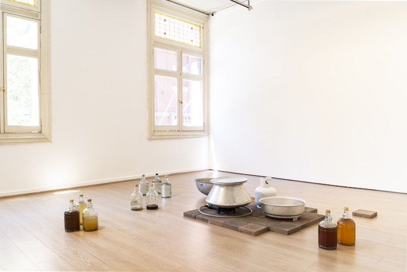 Light coming through a gallery window onto eight glass bottles filled with different liquids, arranged next to an aluminium pot, a two ring gas burner and a gas cylinder on a pavers floor.