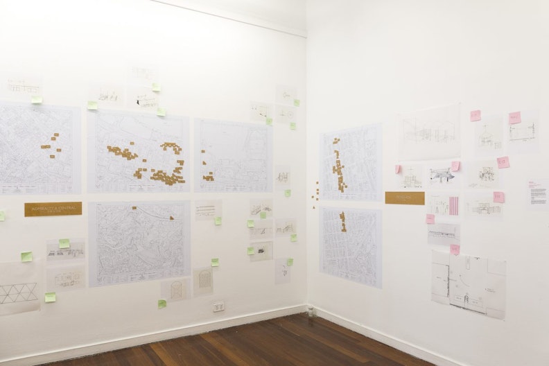 The left and right walls are covered with large printed-maps and architectural drawings, with small brown labels on them, and green and pink memo notes are sticked around.