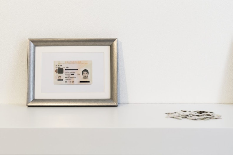 A Hong Kong permanent identity card in a silver frame sits on a white shelf, with a pile of identity card fragments scattered nearby.
